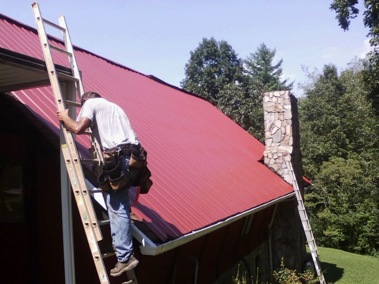 Roofer on a ladder beside a red metal roof and stone chimney — Soffit & Fascia Repair by Roofing Brothers in Trufant